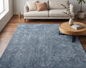 May include: A rectangular area rug in a muted blue tone featuring a grid pattern of small, light-colored cross symbols. The rug has a soft, uniform texture and is positioned on a wooden floor in front of a light-colored sofa and a round table.