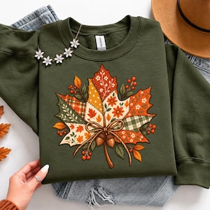 May include: An olive green crewneck sweatshirt featuring a large central graphic of a maple leaf composed of various patchwork patterns in orange, yellow, and green, accented with a tied ribbon, two small acorns, and red berries.
