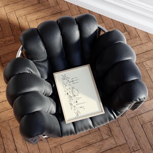 Op de afbeelding: Een zwarte, gecapitonneerde fauteuil met een ingelijste illustratie. De illustratie toont lijntekeningen van bartools, glazen en flessen. De stoel staat op een houten vloer.
