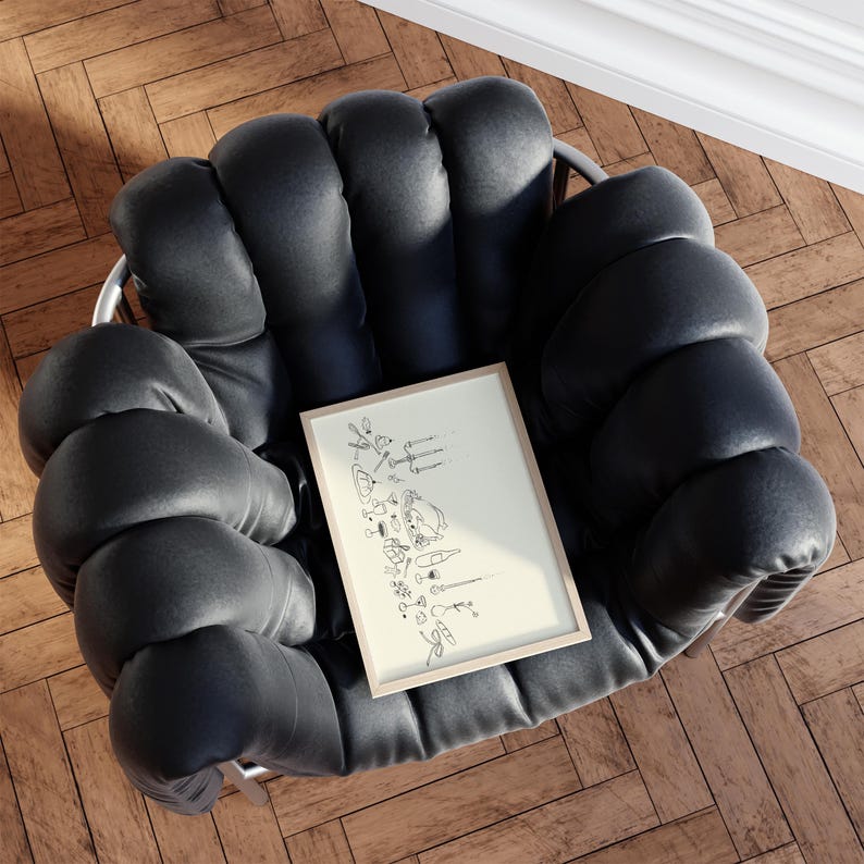 Op de afbeelding: Een zwarte, gecapitonneerde fauteuil met een ingelijste illustratie. De illustratie toont lijntekeningen van bartools, glazen en flessen. De stoel staat op een houten vloer.