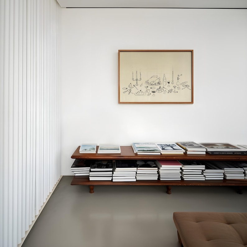 Op de afbeelding: Een kamer met een houten boekenplank vol boeken. Boven de boekenplank hangt een ingelijste tekening. De kamer heeft witte muren en een grijze vloer. Aan de linkerkant van de kamer bevindt zich een witte, verticaal geribbelde wand.