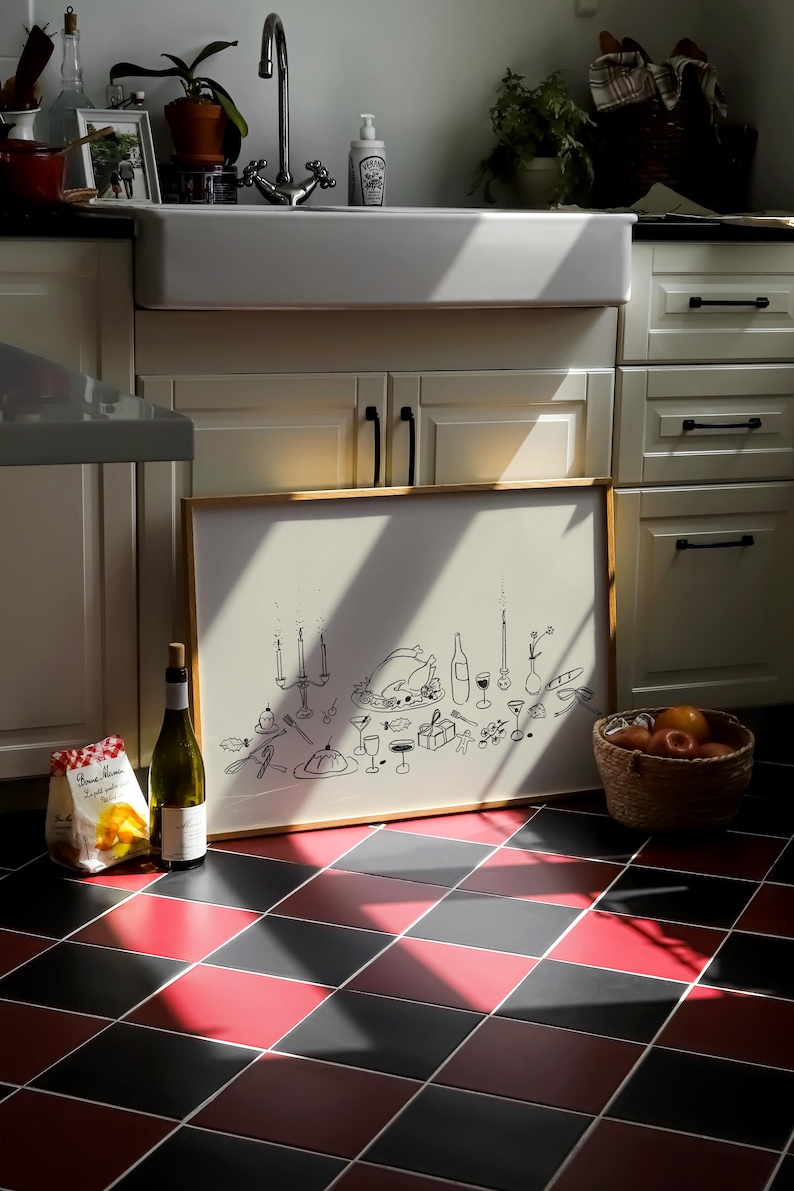 Op de afbeelding: Een ingelijste illustratie van een feestelijk diner, met een kalkoen, kaarsen en wijnglazen. Een fles wijn en een zak pasta staan naast de lijst. Een mand met sinaasappels staat op een rood-zwarte geblokte vloer.