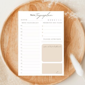 May include: A printable daily planner with a beige background. The planner has a section for daily tasks, a section for priorities, and a section for tracking water intake. The text on the planner reads "Mein Tagesplan", "DATUM", "MEIN TAGESABLAUF", "MDMDFSS", "PRIORITAT DES TAGES", and "WASSER GETRUNKEN".
