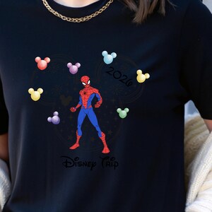May include: Black t-shirt with a Spider-Man design in red and blue, encircled by colourful Mickey Mouse head shapes. The shirt also features the text "Disney Trip 2020" in a circular design.