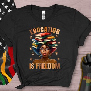 May include: Black t-shirt with the words "EDUCATION IS FREEDOM" in gold lettering. The graphic features a woman with glasses and books stacked on her head. The shirt is next to a Pan-African flag and a black power fist.