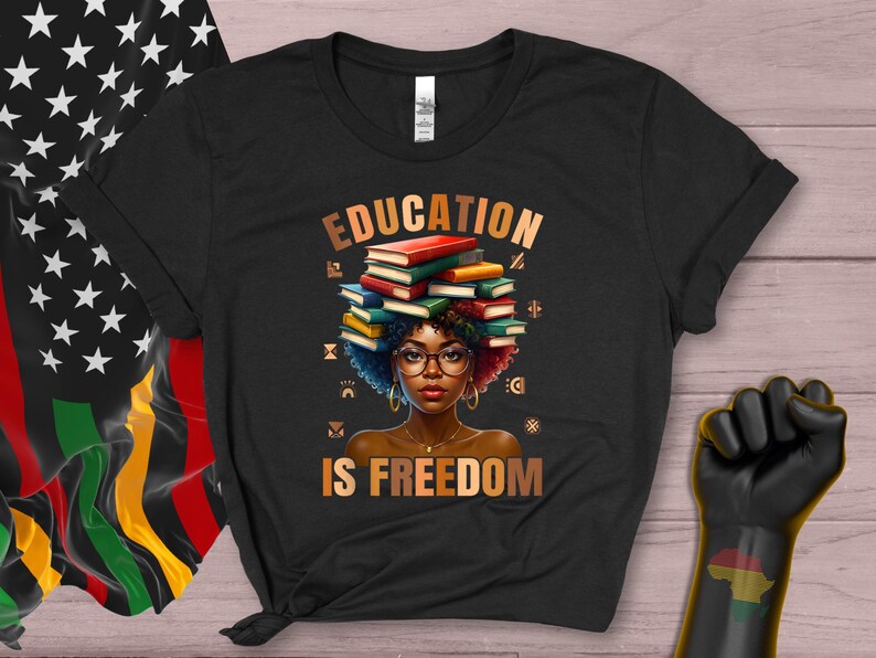 May include: Black t-shirt with the words "EDUCATION IS FREEDOM" in gold lettering. The graphic features a woman with glasses and books stacked on her head. The shirt is next to a Pan-African flag and a black power fist.