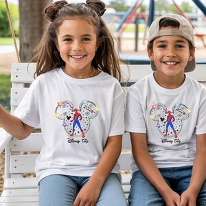 May include: Two children wearing white t-shirts with a Disney-themed graphic. The graphic features a Mickey Mouse head outline with a Spider-Man figure, the year 2024, and the words "Disney Trip". The children are smiling.