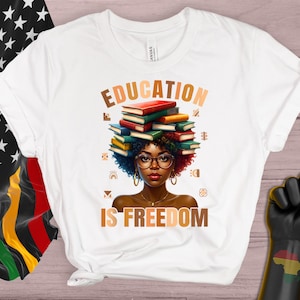 May include: White t-shirt with the words "EDUCATION IS FREEDOM" and an illustration of a person with books on their head. The illustration features a person with glasses and curly hair. A black power fist and a flag with stars and the colors red, black, and green are also visible.