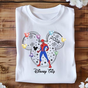 May include: White t-shirt with a graphic of Spider-Man within a Mickey Mouse head outline, with colourful balloons and the text "2026 Disney Trip". The design is in black, red, blue, and yellow.