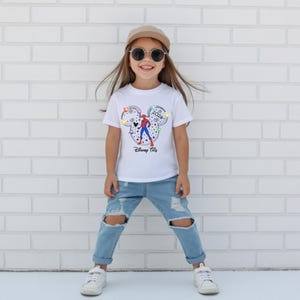 May include: A child wearing a white t-shirt with a Mickey Mouse design featuring a Spider-Man graphic and the text "Disney Trip 2024". The child is wearing blue jeans, sunglasses, a cap, and white trainers.