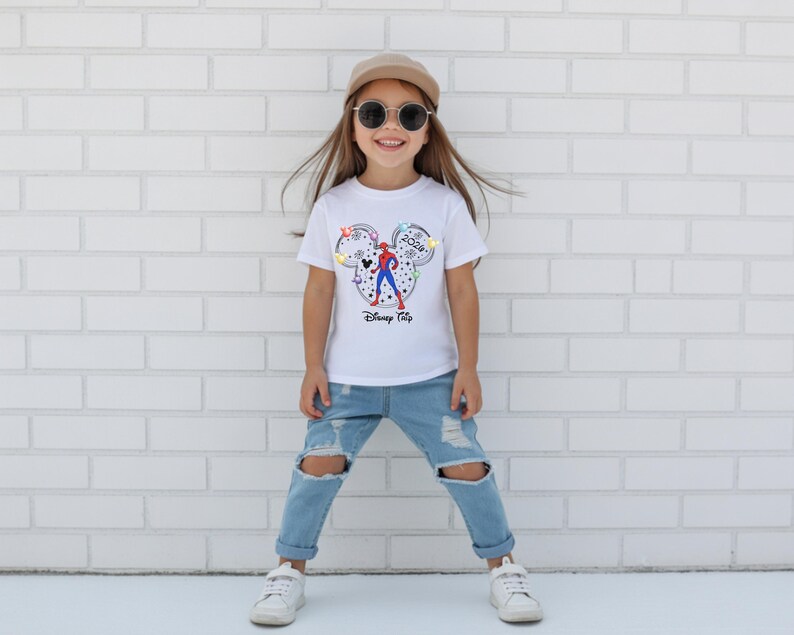 May include: A child wearing a white t-shirt with a Mickey Mouse design featuring a Spider-Man graphic and the text "Disney Trip 2024". The child is wearing blue jeans, sunglasses, a cap, and white trainers.