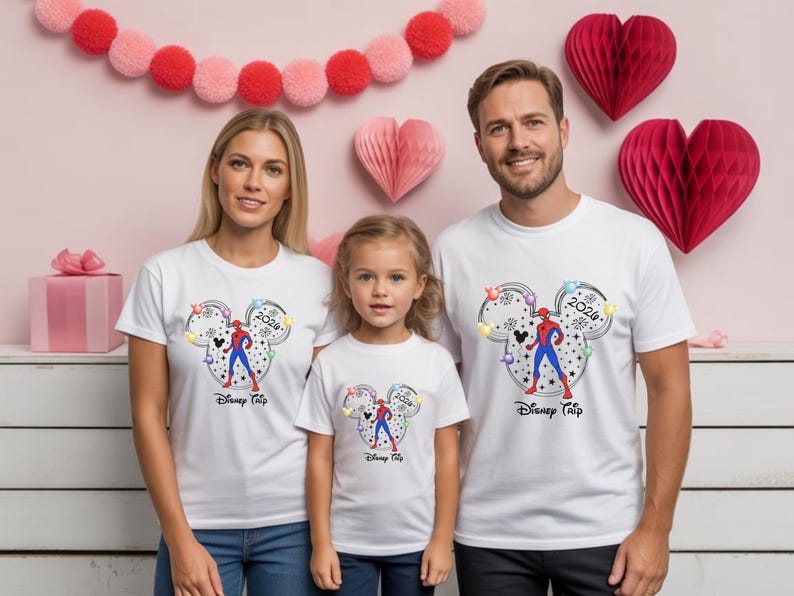 May include: Three people wearing white t-shirts with a Mickey Mouse head design featuring a Spider-Man graphic and the text "Disney Trip 2024". The background includes pink heart decorations and a pink gift box.