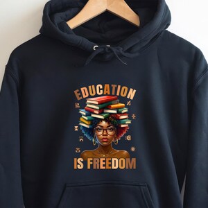May include: Black hooded sweatshirt featuring a graphic of a woman with books stacked on her head, wearing glasses. The text "EDUCATION IS FREEDOM" is displayed in gold lettering. The sweatshirt is hanging on a wooden hanger.