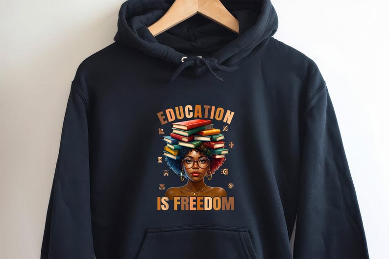 May include: Black hooded sweatshirt featuring a graphic of a woman with books stacked on her head, wearing glasses. The text "EDUCATION IS FREEDOM" is displayed in gold lettering. The sweatshirt is hanging on a wooden hanger.