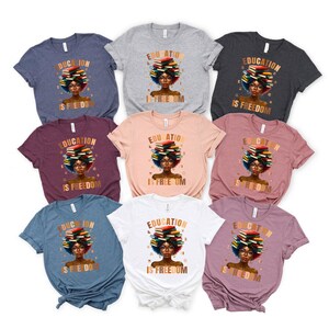 May include: A collection of t-shirts in various colors, each featuring a graphic of a woman with a stack of books on her head. The text "EDUCATION IS FREEDOM" is printed on each shirt. The shirts are short-sleeved.