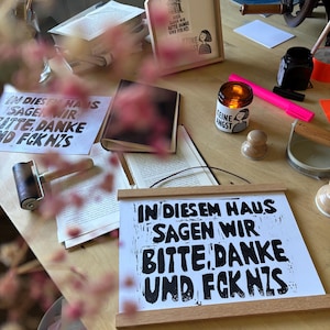 Könnte beinhalten: Ein weißes rechteckiges Poster mit schwarzem Aufdruck IN DIESEM HAUS SAGEN WIR BITTE, DANKE UND FCK NZS, das oben und unten von zwei horizontalen Holzleisten gehalten wird. Das Poster liegt auf einer Holzoberfläche neben einer Kerze und Büchern.