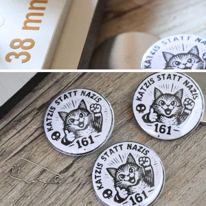Puede incluir: Tres chapas circulares blancas con ilustraciones en tinta negra de un gato haciendo un gesto de pulgar hacia arriba. El texto dice "KATZIS STATT NAZIS" y "161" con un pequeño icono de calavera. Se ve un imperdible y una regla de 38 mm.