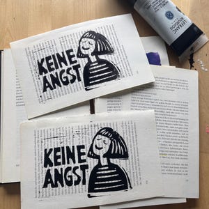 Könnte beinhalten: Zwei rechteckige Papierdrucke mit einem schwarzen Linoldruck einer Person mit Bob-Frisur und gestreiftem Oberteil. Der Text KEINE ANGST ist in fetten schwarzen Buchstaben neben der Figur auf Buchseiten gedruckt.