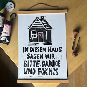 Könnte beinhalten: Ein weißes Poster mit einer schwarzen Linoldruck-Illustration eines Hauses und einem kleinen rosa Herz an der Tür. Der Text lautet: IN DIESEM HAUS SAGEN WIR BITTE, DANKE UND FCK NZS. Das Poster wird von zwei magnetischen Leisten aus hellem Holz gehalten.