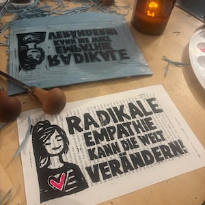 May include: A rectangular linocut block with a carved mirror-image design and a corresponding paper print featuring a stylized illustration of a person holding a heart with the German text RADIKALE EMPATHIE KANN DIE WELT VERÄNDERN printed in black ink.