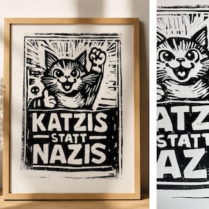 Könnte beinhalten: Ein gerahmter schwarz-weißer Linoldruck mit einer stilisierten Katze, die eine Faust hebt. Die Katze hat große Augen und ein breites Lächeln. Unter der Katze steht der Text KATZIS STATT NAZIS in fetten, blockartigen Großbuchstaben. Heller Holzrahmen.