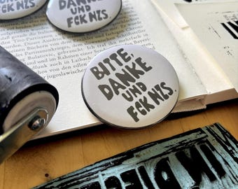 Könnte beinhalten: Drei runde weiße Ansteckbuttons mit dem schwarzen Text BITTE, DANKE UND FCK NZS. Die Buttons liegen auf einer Fläche neben einer schwarzen Farbwalze und einem geschnitzten Druckstock mit gleichem Text sowie einer bedruckten Buchseite.