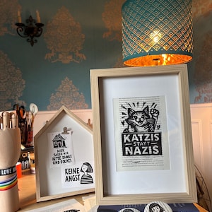 May include: A light wood frame containing a black and white print of a cat with the text KATZIS STATT NAZIS. Next to it is a house-shaped display board with two small prints, one showing a house and text, the other a person and text KEINE ANGST.