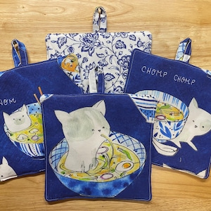 May include: A collection of five square pot holders. Four are navy blue with white cat illustrations in ramen bowls, and the text "Nom Nom" and "Chomp Chomp". The fifth is white with a blue floral pattern.