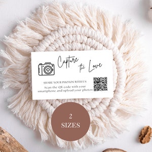 May include: White card with black text that says "Capture the Love" and a camera icon with a heart inside. Below the text is a QR code and the text "SHARE YOUR PHOTOS WITH US Scan the QR code with your smartphone and upload your photos". The card is sitting on a white macrame circle with brown fringe. Below the card is a brown circle with the text "2 SIZES" in white.