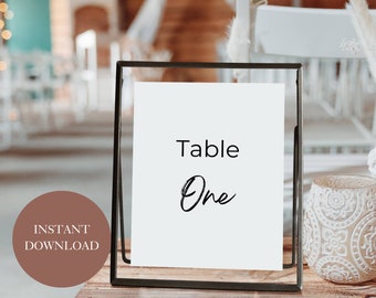 Könnte beinhalten: Ein schwarzer Metallrahmen mit einer weißen Karte darin. Die Karte trägt den Text "Table One" in schwarzer Kalligraphie.