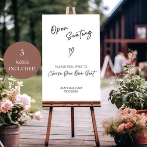May include: White sign with black calligraphy text that reads "Open Seating" with a heart symbol. The text below reads "Please feel free to choose your own seat." The sign also includes the names Amelia & Liam and the date 19.10.2024. The sign is on an easel in front of a wooden building.