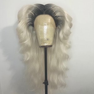 Könnte beinhalten: Eine lange, blonde Perücke mit dunklen Wurzeln wird auf einem Schaufensterkopf präsentiert. Die Perücke hat voluminöse, wellige Schichten und eine Lace-Front. Der Schaufensterkopf ist mit einem beigefarbenen Stoff bedeckt und auf einem schwarzen Ständer montiert.