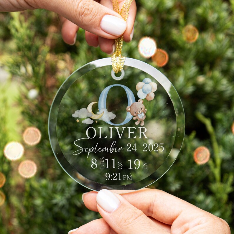 Pu&ograve; includere: Ornamento rotondo trasparente con nastro dorato. L'ornamento presenta il nome "OLIVER", la data "24 settembre 2025" e i dettagli della nascita. Elementi decorativi: orsacchiotto, palloncini, luna e stelle. I dettagli includono il peso in kg e g.