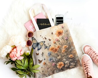 May include: A white canvas tote bag featuring a watercolour design of butterflies and flowers. The bag is filled with a pink notebook, a pink book, a white phone, and sunglasses.