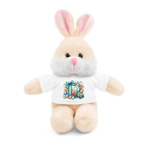Op de afbeelding: Een pluche witte bunny die een wit T-shirt draagt met een blauw en groen paasei-ontwerp. Het ontwerp toont een bunny die een mand met eieren in een vrachtwagen vervoert.