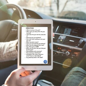 Op de afbeelding: Een witte tablet met een blauwe homeknop toont een lijst met vragen over buikpijn. De tablet wordt vastgehouden door een persoon in een auto.