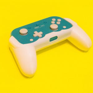 Pode incluir: Um controle de jogo sem fio branco e verde-azulado com um direcional em forma de cruz e vários botões. O controle tem um design arredondado e está sobre um fundo amarelo brilhante.