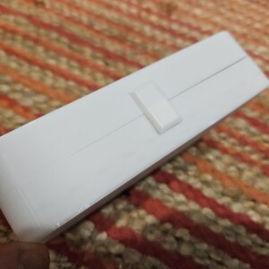 May include: A white rectangular box with a small, rectangular clasp. The box is on a patterned surface with orange, brown, and beige stripes. The box is held by a person's fingers.