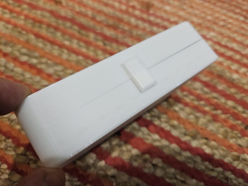 May include: A white rectangular box with a small, rectangular clasp. The box is on a patterned surface with orange, brown, and beige stripes. The box is held by a person's fingers.