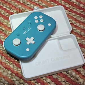 Pode incluir: Um controle de jogo 8BitDo Lite 2 azul-turquesa com botões brancos e uma caixa branca. A caixa está aberta, revelando o controle e o texto "DKINT Customs" na caixa. O controle está em uma superfície tecida multicolorida.