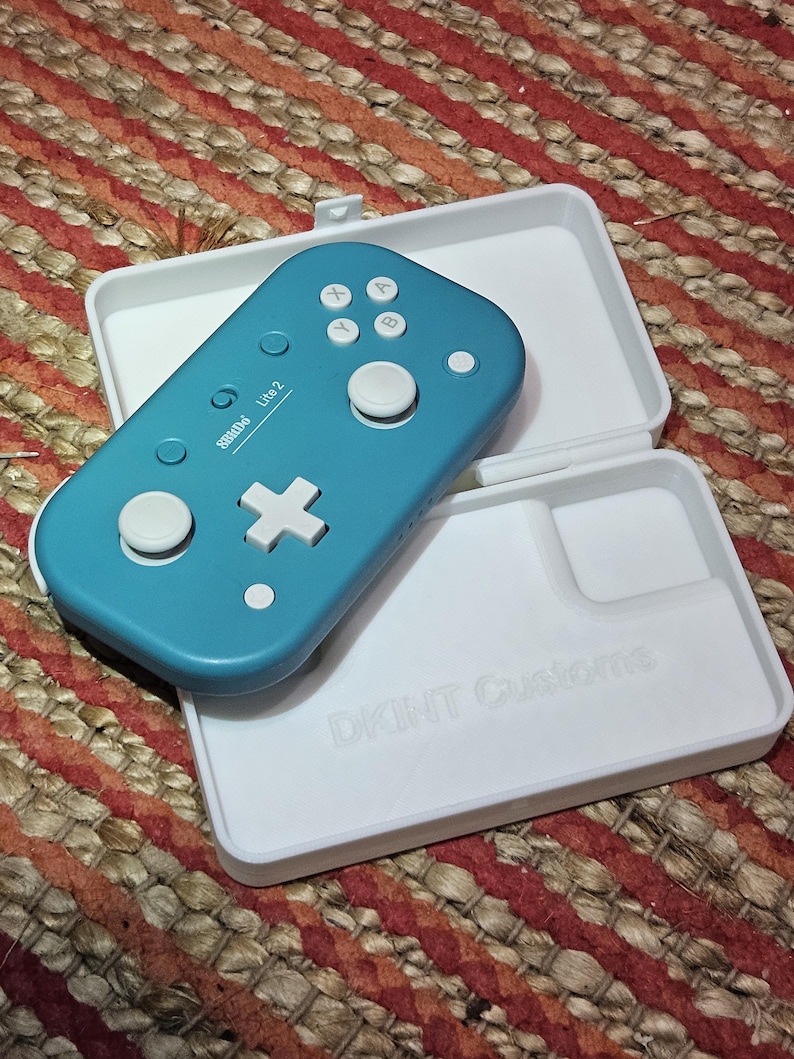 May include: A teal-colored 8BitDo Lite 2 game controller with white buttons and a white case. The case is open, revealing the controller and the text "DKINT Customs" on the case. The controller is on a woven, multi-colored surface.