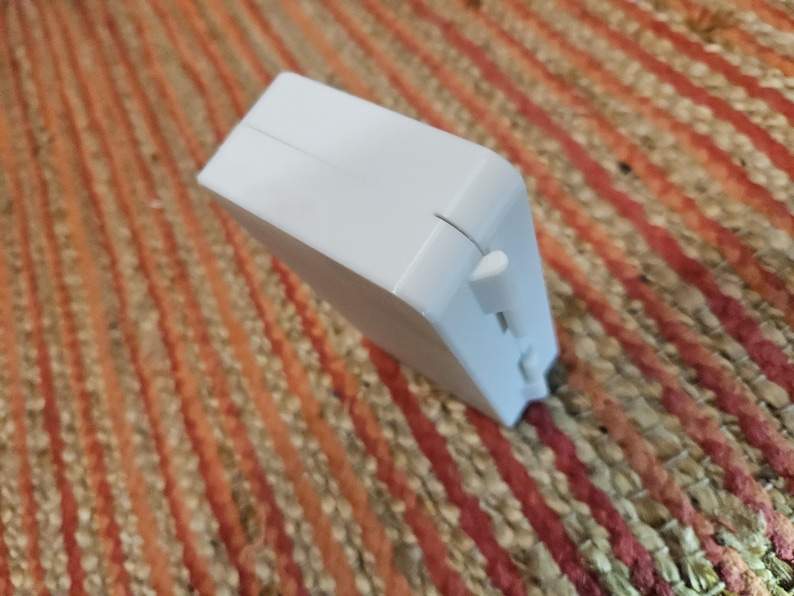 May include: A white rectangular object with a small, protruding button on one side. The object is angled, revealing a side with a small opening. The background is a textured surface with orange and red stripes.