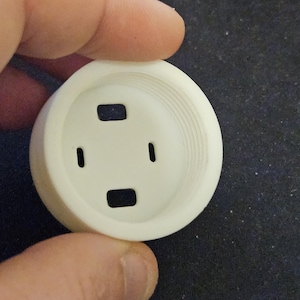 May include: A white, circular component featuring four rectangular apertures and a threaded internal structure. The object is held in a hand, highlighting its design. It appears to be a part of a larger product.