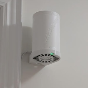 May include: A white air purifier mounted on a wall. The cylindrical device has a fan-like base and a series of diagonal lines on the side. A green light is illuminated on the base. The device is attached to the wall with a white bracket.