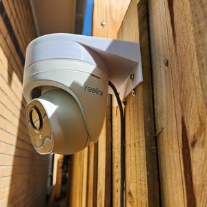 Puede incluir: Una cámara de seguridad blanca con la marca "reolink" está montada en una valla de madera. La cámara tiene un diseño redondeado y está sujeta a un soporte blanco. Un cable negro sale de la cámara.