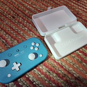May include: A teal SBIDD Lite 2 controller with white buttons and a white directional pad, next to an open white case with the text "DKINT Customs". The case is designed to hold the controller. The items are on a brown and orange woven surface.