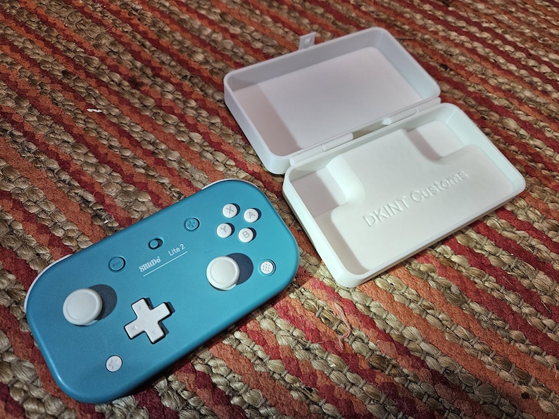 May include: A teal SBIDD Lite 2 controller with white buttons and a white directional pad, next to an open white case with the text "DKINT Customs". The case is designed to hold the controller. The items are on a brown and orange woven surface.