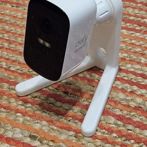 Op de afbeelding: Een witte Eufy beveiligingscamera met een zwarte lens en het woord "eufy SECURITY" gedrukt op de zijkant. De camera is gemonteerd op een witte standaard met twee poten. De camera staat op een geweven tapijt.