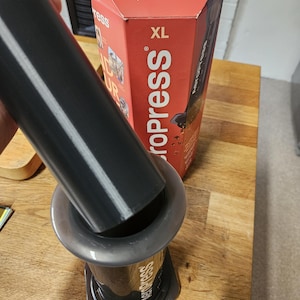 Puede incluir: Cafetera AeroPress XL con un émbolo negro y una cámara gris. La caja roja y blanca dice "AeroPress XL" y "Find Your Inner Brew". La cafetera está sobre una superficie de madera.