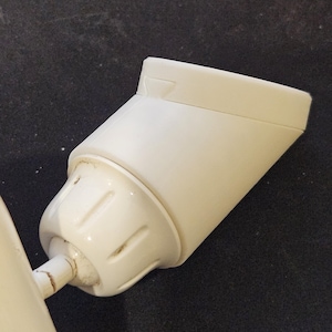 May include: A close-up of a white, angled light fixture component. The fixture has a rectangular top and a rounded base, connected by a cylindrical arm. The surface shows slight wear and tear. The background is a dark, textured surface.